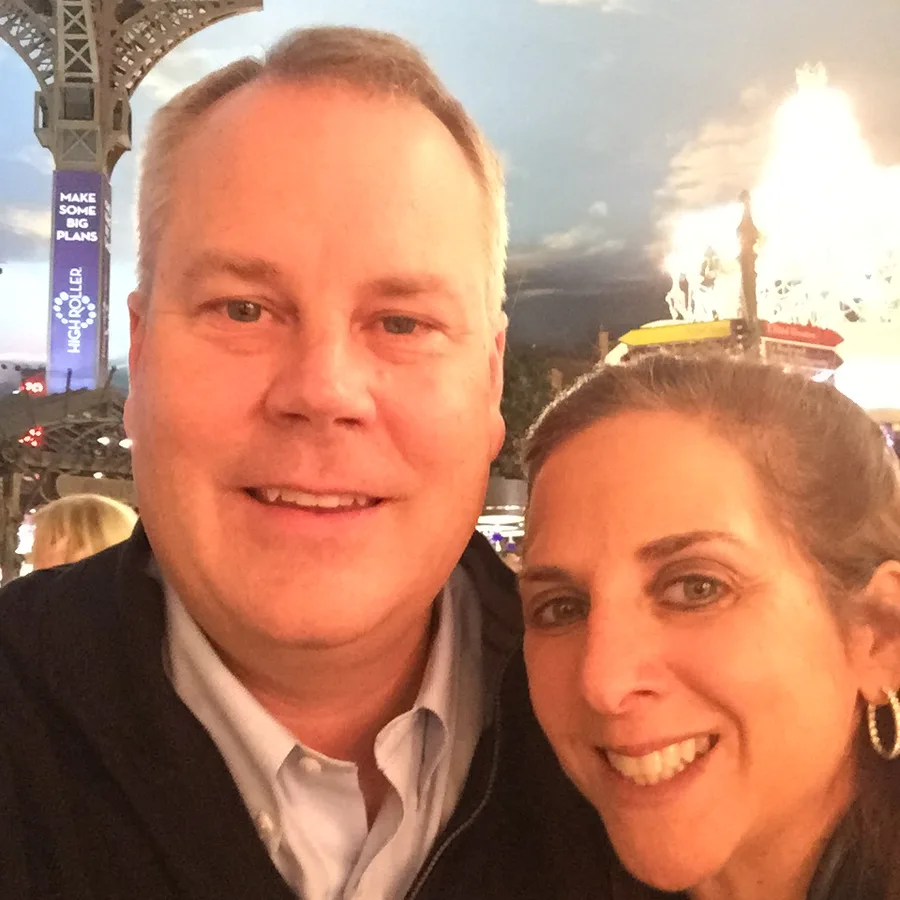 A man and a woman cheek to cheek at night, a brightly lit fairground behind them, both smiling.